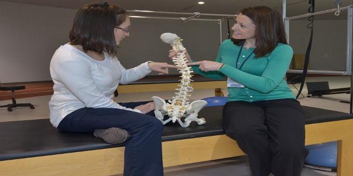 一个治疗师和一个病人坐在一张垫子桌上讨论脊柱的解剖，中间有一个模型.